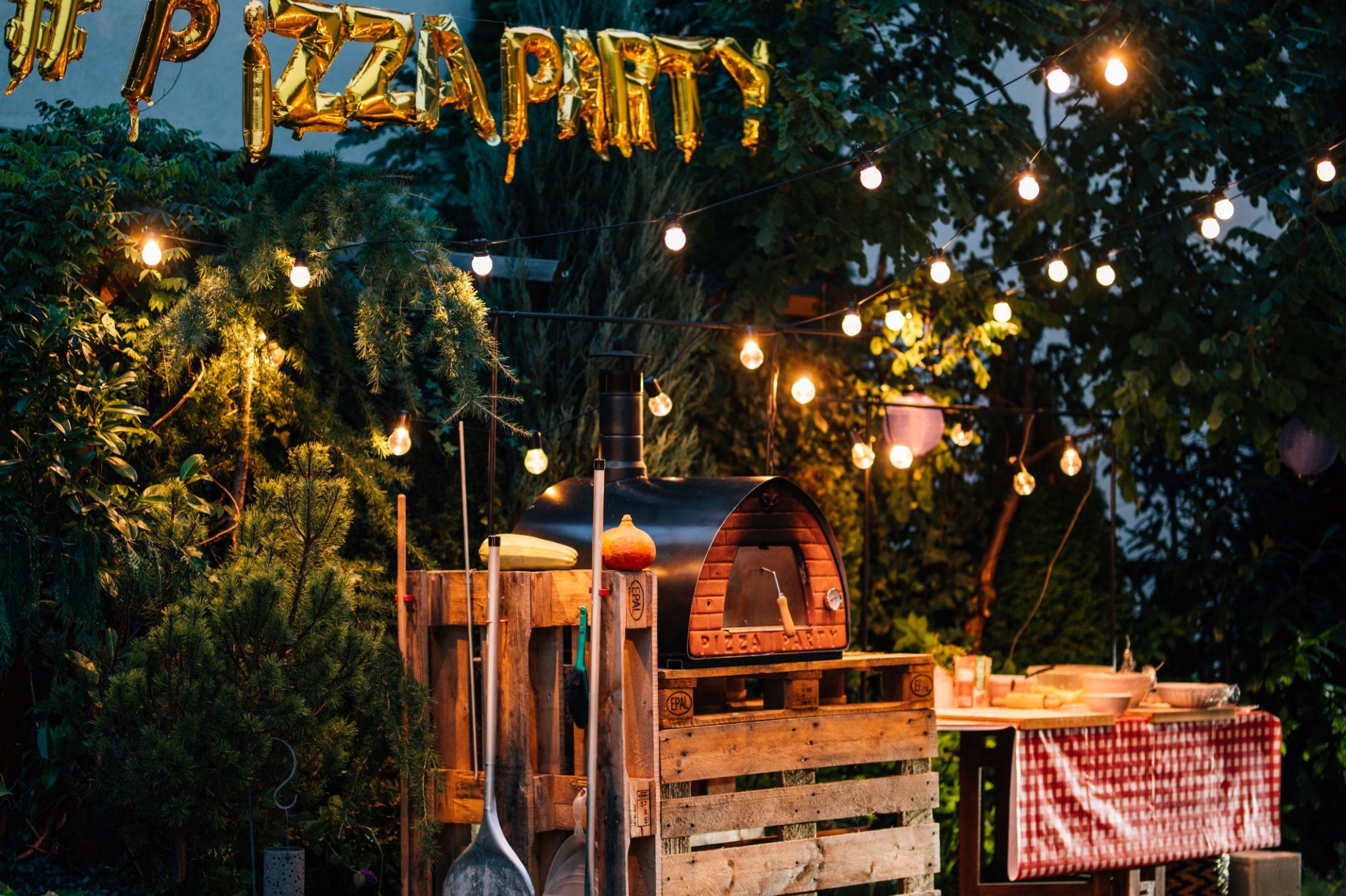 Ogrodowy piec do pizzy Pizza Party opalany drewnem w klimatycznej aranżacji z girlandami świetlnymi.