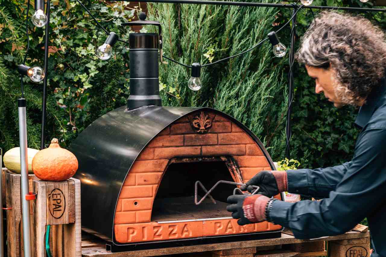 Osoba obsługująca piec do pizzy Pizza Party opalany drewnem w ogrodowej przestrzeni z dekoracyjnym oświetleniem.